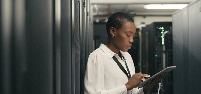 A Black woman in professional clothing interacts with a tablet she is holding inside a server room