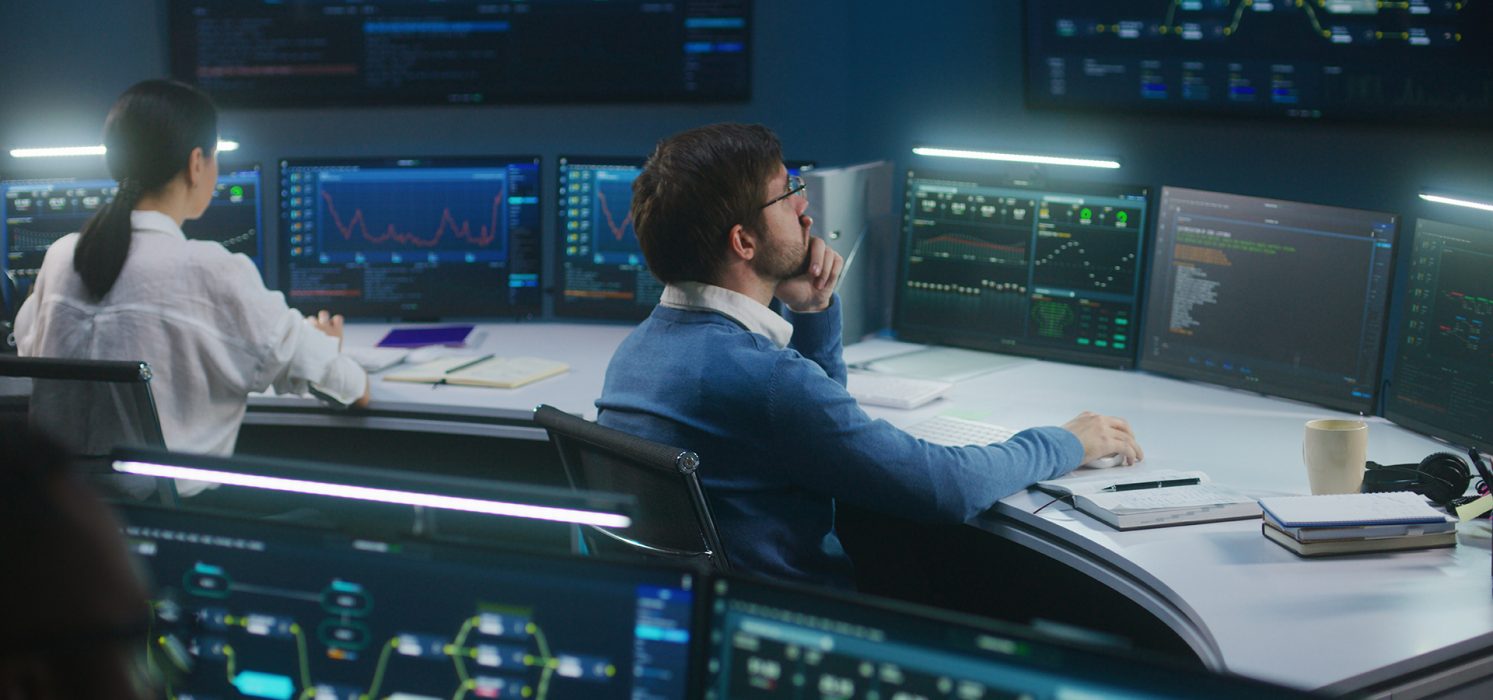 Two people in business attire sit in an office environment, working in front of multiple monitors at each station. The screens display charts and data.