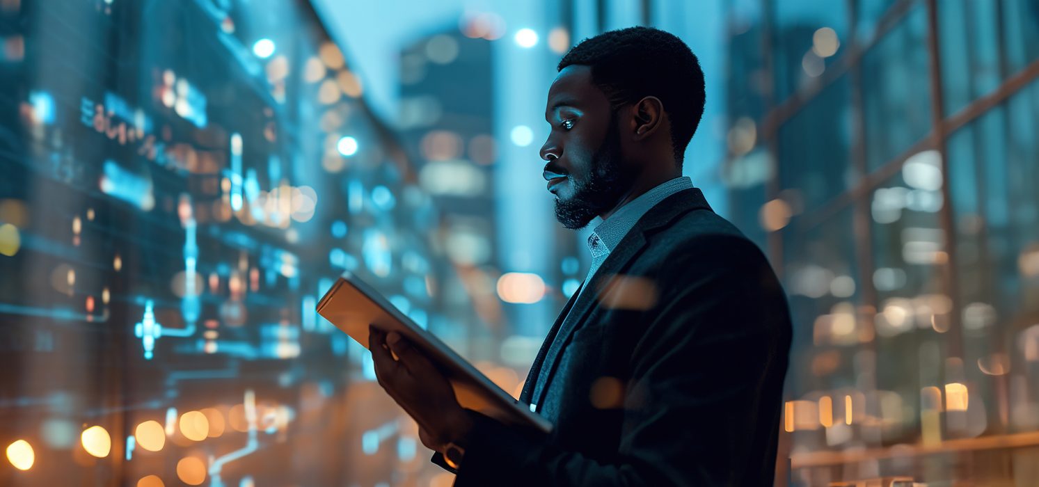A Black man in business attire using a tablet, with illuminated skyscrapers in the background.
