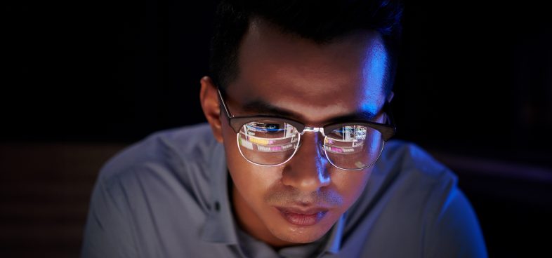 A person focuses intently on a screen, with many lines of code on the monitor reflected in their glasses.