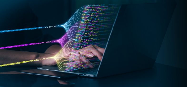 A person's hands type on a laptop. Superimposed graphically before the screen is code appearing in three separate streams by color, before joining together in many lines.