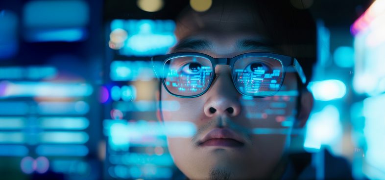 An Asian man wearing glasses sits in front of a computer screen. Reflecting in the glasses are lines indicating analysis. Bright blue city lights illuminate the rest of the image.