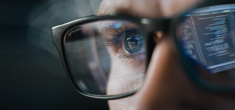 Pictorial representation of attack surface management. Close-up of a person wearing glasses with computer code reflections on the lenses.