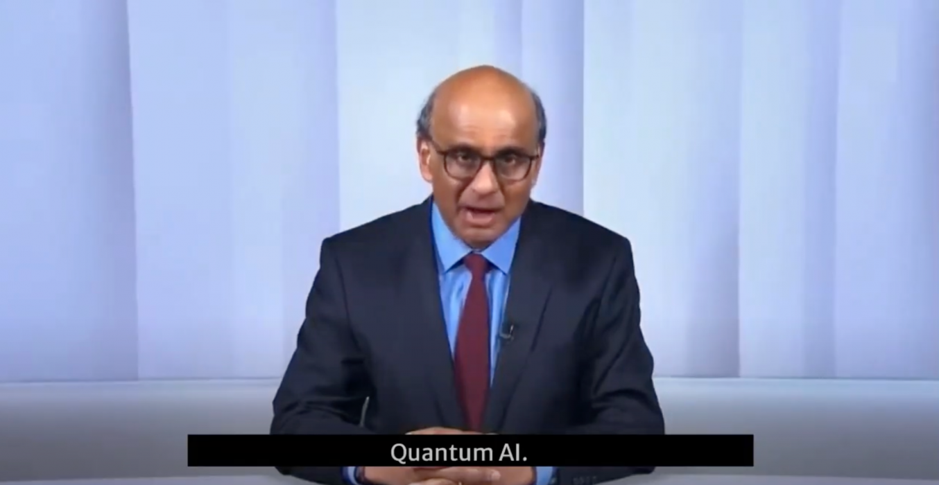 A person in business attire is seated at a news desk with the words "Quantum AI" displayed below. The background features a plain, light-colored wall.