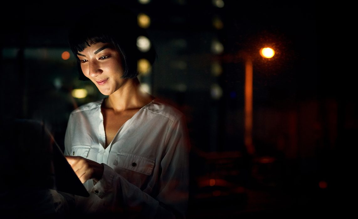 Pictorial representation of a female smiling while looking at the screen of a tablet. The backgorund is dark and features warm-toned lights.